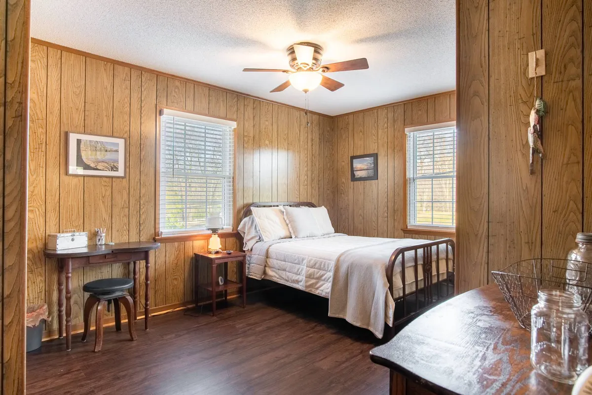 A tranquil bedroom with a comfortable bed, a small table and chairs, large windows letting in natural light, and a ceiling fan overhead. The space feels airy and functional, suitable for both relaxation and quiet activities like reading or writing.