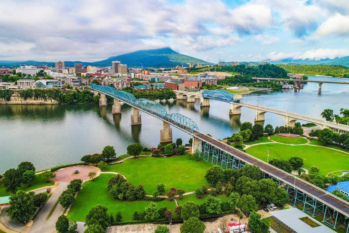 A sweeping view of the Chattanooga city skyline with the prominent blue bridge, the river glistening below, and Lookout Mountain in the background. The scene is dynamic, capturing walkers and joggers along the riverside park as the morning sun lights up the city.