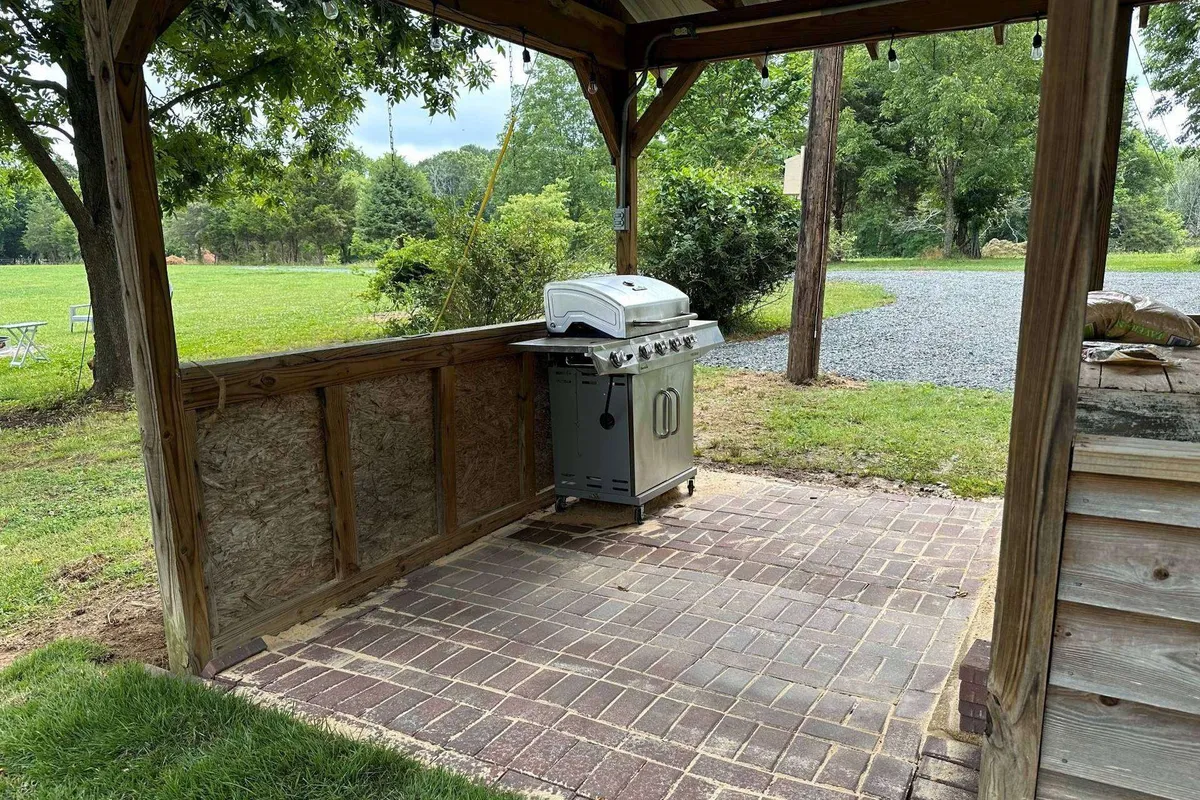 An exterior view with a grill, a wooden gazebo-style structure, and surrounding greenery. The scene highlights a relaxed atmosphere for outdoor cooking and dining, with space for groups to gather and enjoy fresh air meals.