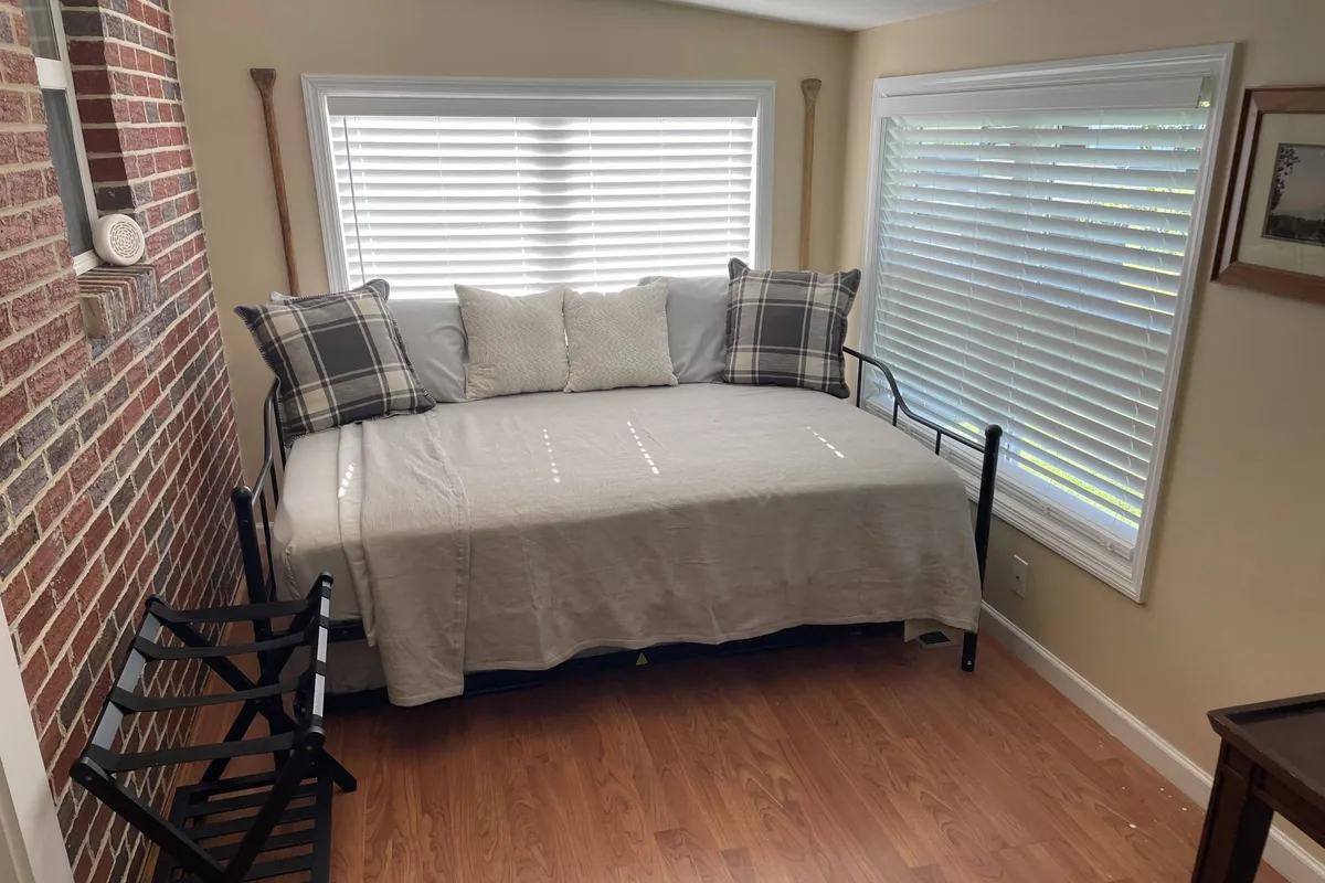 A sunlit daybed with plaid pillows sits beside a window, with a wooden paddle as wall decor and a brick wall adding texture. The space feels playful and inviting—a great spot for lounging, reading, or accommodating extra guests.