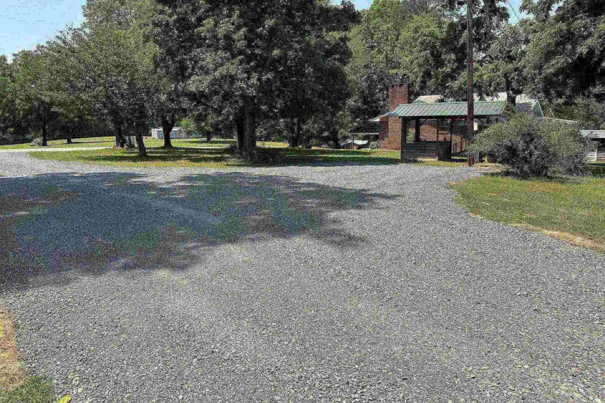 A wide gravel driveway bordered by lush trees, leading to a small structure with a chimney, illustrating plentiful parking and easy access for vehicles, boats, or trailers.