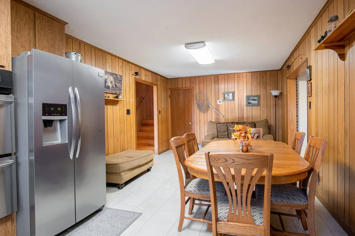 An inviting interior featuring a dining table, refrigerator, comfortable sofa, and wood paneling. The arrangement encourages group meals, board games, or relaxed conversation, all set in a warm, communal atmosphere.
