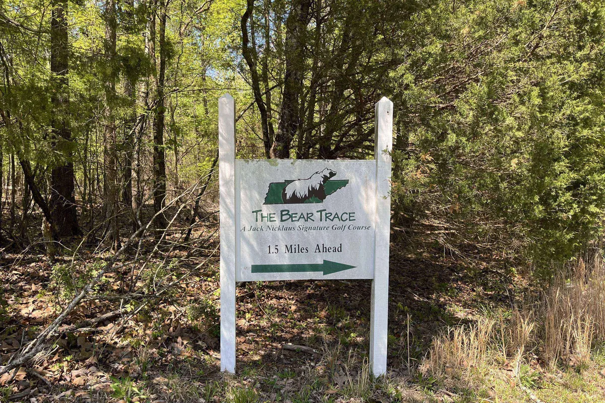 The image features the sign for The Bear Trace golf course, surrounded by lush greenery, with a road leading further into the scenic area. The entrance is inviting and hints at the vibrant golfing experience that awaits just steps away from the property.
