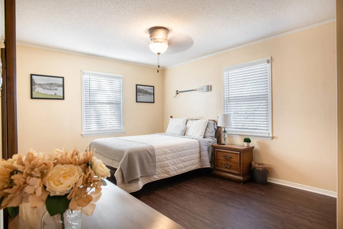 A serene bedroom scene with a neatly made bed, a nightstand adorned with a flower arrangement, and charming decorative photos on the wall. Soft daylight filters through a nearby window, creating a calm, cozy atmosphere ideal for relaxation and restful sleep.