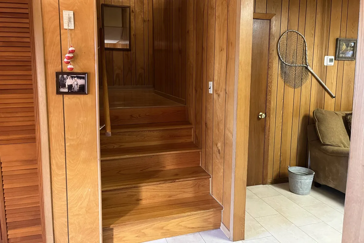 A warm, homey room with a wooden staircase, a cozy couch, a framed photo on the wall, and a racket for a playful touch. The space suggests relaxing, storytelling, and family gatherings, with a lived-in feel that encourages sociability.