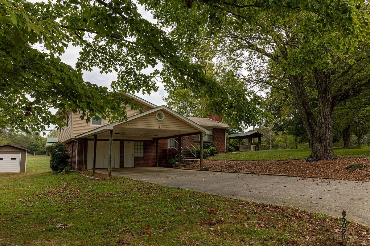 A welcoming exterior view featuring the front porch of the property, a large tree for shade, garage, and a meticulously landscaped yard. The space is clearly designed for gatherings, relaxation, and outdoor activities, with plenty of room for parking boats and trailers.