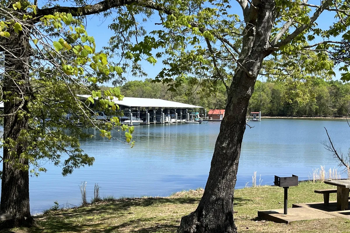 A panoramic lakeside view with a tranquil lake, a boathouse, trees, and a picnic area along the shore. The image suggests options for boating, fishing, and picnicking, with calm water reflecting the sky and greenery.