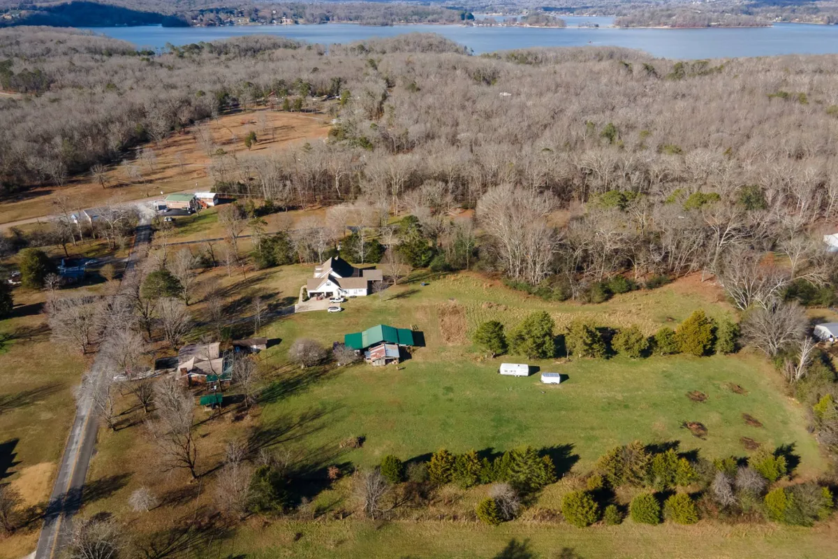 The image showcases a spacious green-roofed family house nestled among mature trees, with a wide, lush lawn stretching toward a sparkling lake in the background. The peaceful setting suggests ample space for outdoor activities, family gatherings, and enjoying nature, while the water and forested parkland hint at nearby recreational opportunities.
