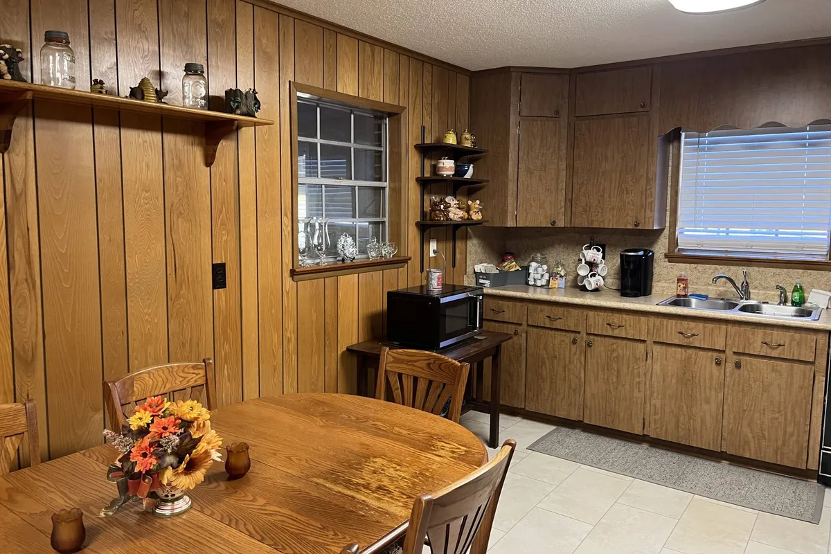 An inviting interior featuring a wooden dining table set for a group, shelves with decorative items, and a kitchen countertop with visible appliances. The space is clearly designed for shared meals and conversation, with seamless flow between kitchen and dining areas.